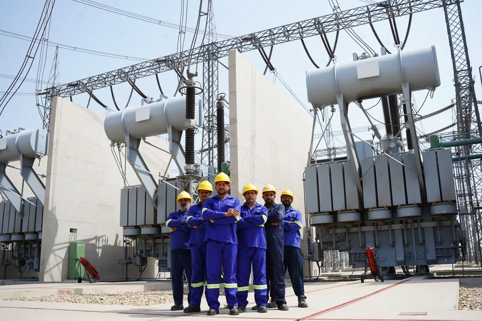 Six National Grid personnel in blue workwear and yellow hard hats standing in front of substation equipment
